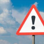 Warning sign with exclamation mark against cloudy sky.