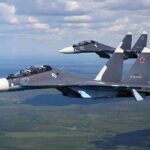 Two fighter jets flying in formation over green landscape.