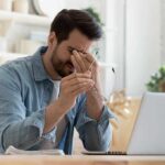 Man holding glasses, looking tired at laptop.