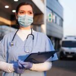Nurse in scrubs and mask outside hospital holding clipboard