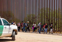 People walking beside tall fence and border patrol vehicle