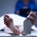 A deceased body on a table with a tag on the foot, and a medical professional in the background