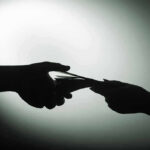 Border Czar OUSTED In Bribery Shocker – Undercover FBI Sting! Silhouette of hands exchanging money in dim light.