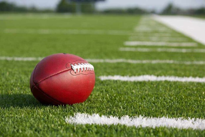 A red American football resting on a green artificial turf field