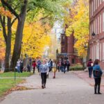 Students walking on a campus path surrounded by autumn trees and brick buildings