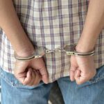 A person in handcuffs viewed from behind, wearing a plaid shirt and denim jeans