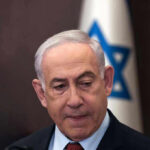 Man in suit with Israeli flag in background.