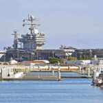 Hegseth DEPLOYS Largest U.S Warship – See Where It’s Headed A large naval aircraft carrier docked in a harbor with smaller boats in the foreground