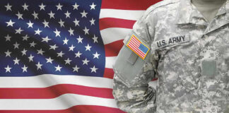 Soldier in camouflage uniform standing before American flag.