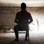 Person tied to chair in dimly lit room.