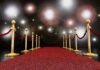 A red carpet lined with gold stanchions and ropes under bright spotlights
