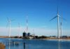 Wind turbines and a communication tower near a body of water under a clear blue sky