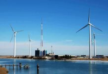 Wind turbines and a communication tower near a body of water under a clear blue sky