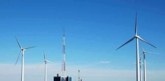 Wind turbines and a communication tower near a body of water under a clear blue sky