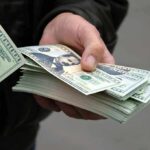 Britain Abolished Slavery — Now FORCED to Pay A hand holding a stack of hundred dollar bills with one bill prominently displayed
