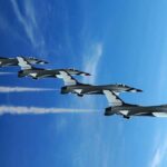 Four military jets flying in formation against a blue sky, leaving smoke trails