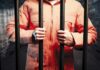 Person in an orange jumpsuit gripping prison bars in a dark cell