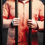 Person in an orange jumpsuit gripping prison bars in a dark cell