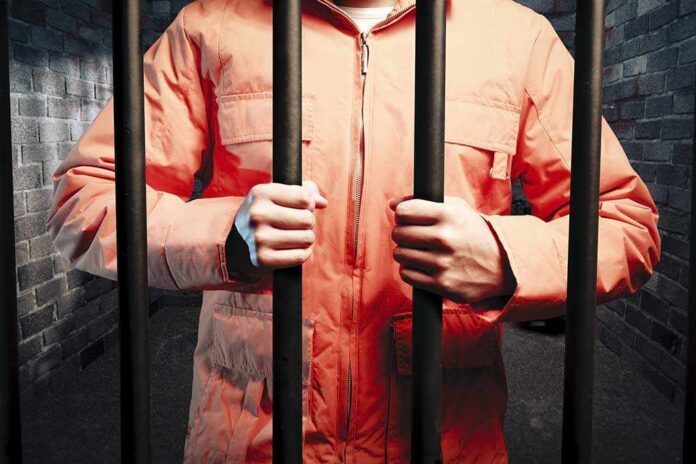 Person in an orange jumpsuit gripping prison bars in a dark cell