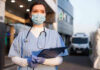 Cardiac Arrest Dispatch Raises Fresh Questions Nurse in scrubs and mask outside hospital holding clipboard.