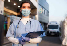 Cardiac Arrest Dispatch Raises Fresh Questions Nurse in scrubs and mask outside hospital holding clipboard.