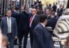 Political Rant Ends in Viral Crash Man in suit waves while surrounded by security detail on a city street
