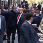 Man in suit waves while surrounded by security detail on a city street
