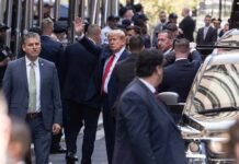Man in suit waves while surrounded by security detail on a city street