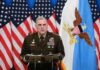 Military officer speaks at podium with U.S. flags behind