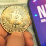 Person holding Bitcoin coin, NFT text on screen.