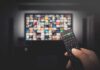 Hand holding TV remote toward a screen with streaming thumbnails