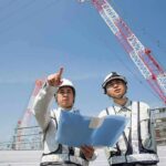 Two construction workers reviewing plans at a site with cranes