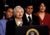 Federico Peña stands behind a woman speaking at a podium with the presidential seal