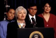 Federico Peña stands behind a woman speaking at a podium with the presidential seal