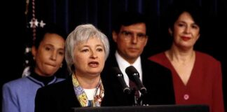 Federico Peña stands behind a woman speaking at a podium with the presidential seal