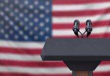 Podium with microphones in front of a blurred American flag