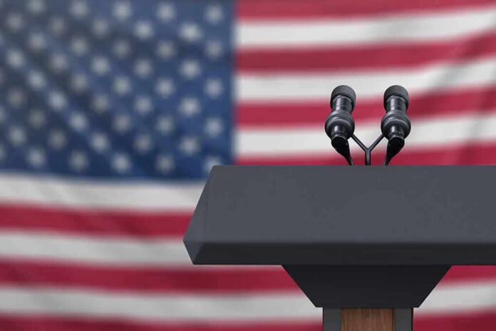 shutterstock_1223942518 Podium with microphones in front of a blurred American flag