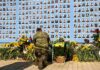 Soldier kneels at memorial wall with photos and flowers