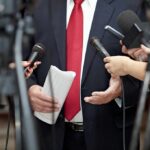 Trump Pays Tribute to Bears Legend Man in suit with red tie speaking to reporters holding microphones