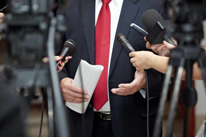 shutterstock_82082848 Man in suit with red tie speaking to reporters holding microphones